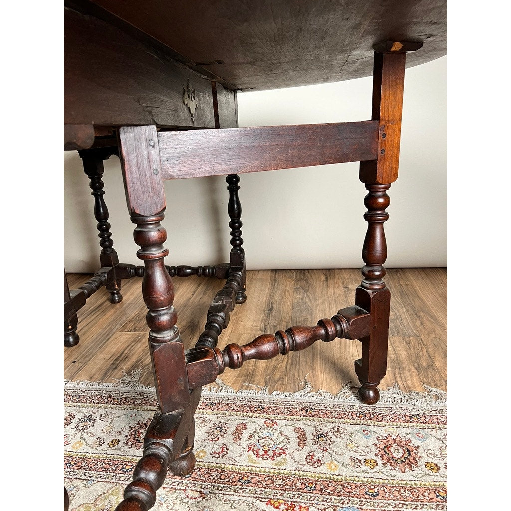 Close-up of the legs of an Antique English William & Mary Walnut One Drawer Gateleg Dining Table (early 18th century), featuring ornate turned details, standing on a patterned rug over a wooden floor against a plain light wall.