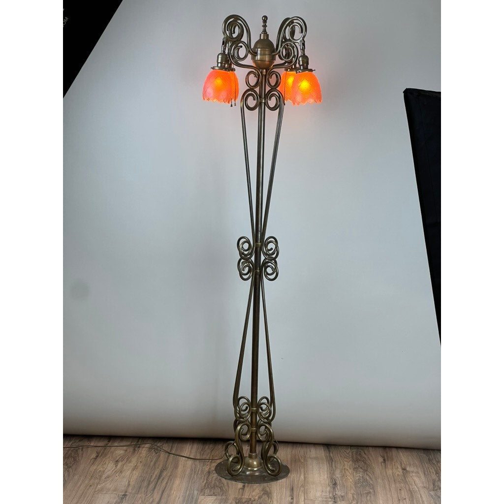 An antique Art Nouveau brass floor lamp, attributed to Jules Cayette and Charles Schneider c. 1920, features intricate scrollwork and three glowing orange-red glass lampshades, standing on a wooden floor against a light backdrop. Brand: Unknown.