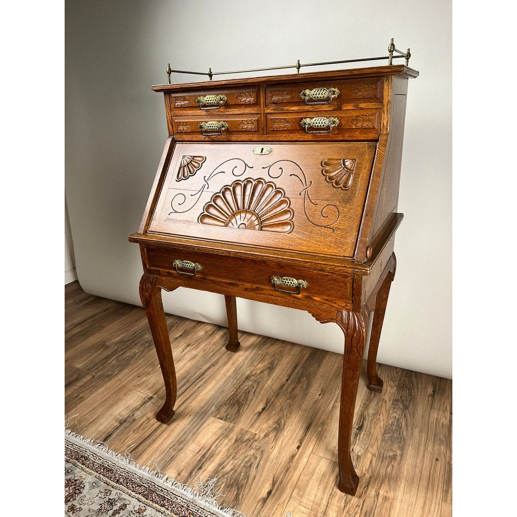 The Antique Victorian Oak Ladies Writing Desk c. 1900 features curved legs, ornate brass handles, carved floral and shell front details, and a drop-down writing surface, shown on a wood floor next to a patterned rug. Brand: None/Unknown.