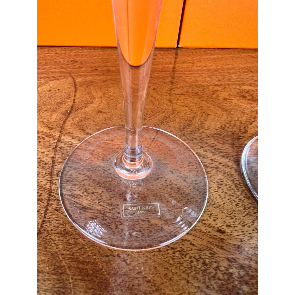 A close-up of the base of a Hermès Saint Louis France crystal champagne flute from a set of eight 10.5’’ flutes, shown on a wooden surface with the label visible and an orange background highlighting this luxury collectible glassware.