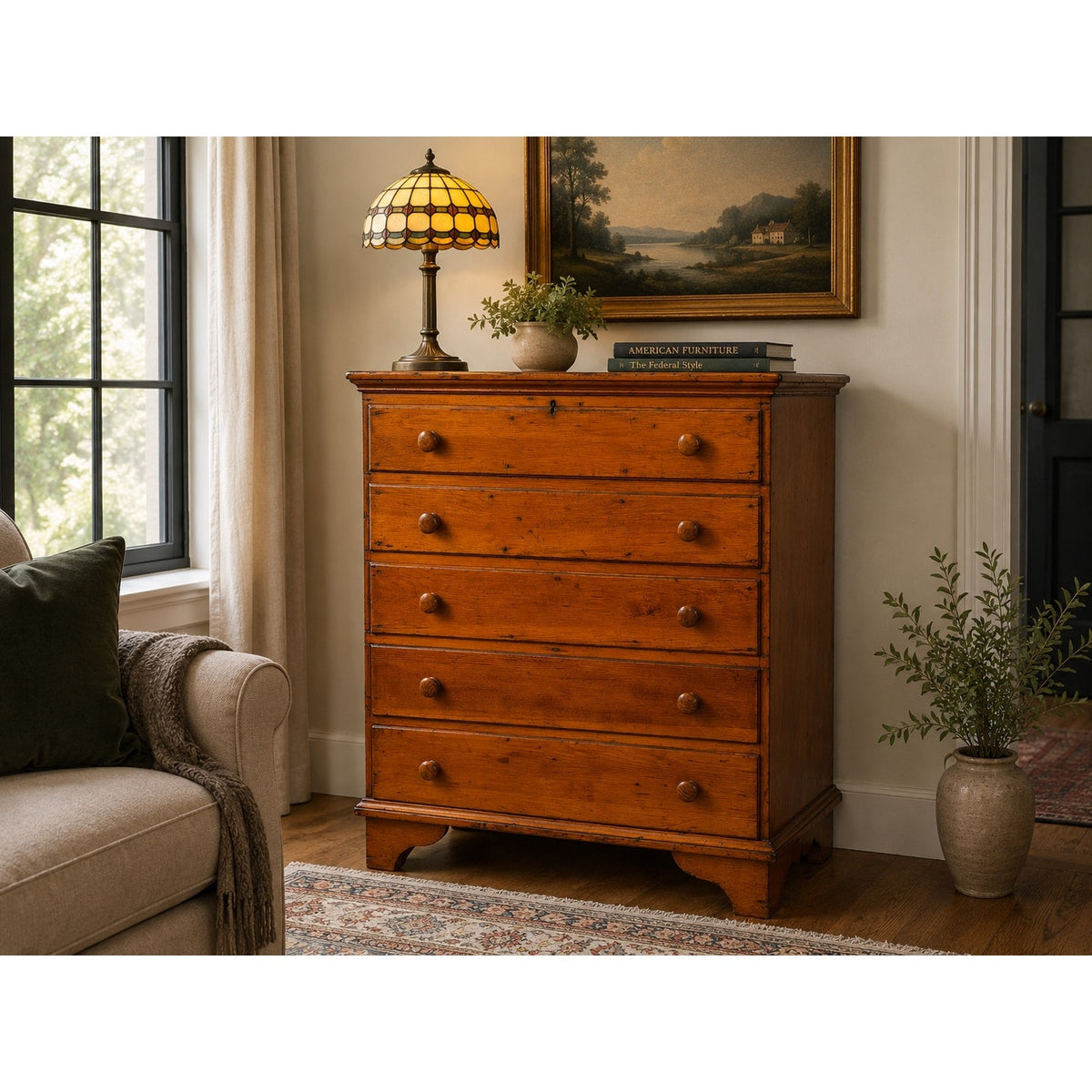 A Late 18th Century New England Pine Two-Drawer Blanket Chest by Pearl City Antiques, dating from around 1780–1810, stands against the wall in a cozy living room with a couch, potted plant, large windows, and a framed painting. The chest has a lamp and books on top, evoking the charm of early American country furniture.