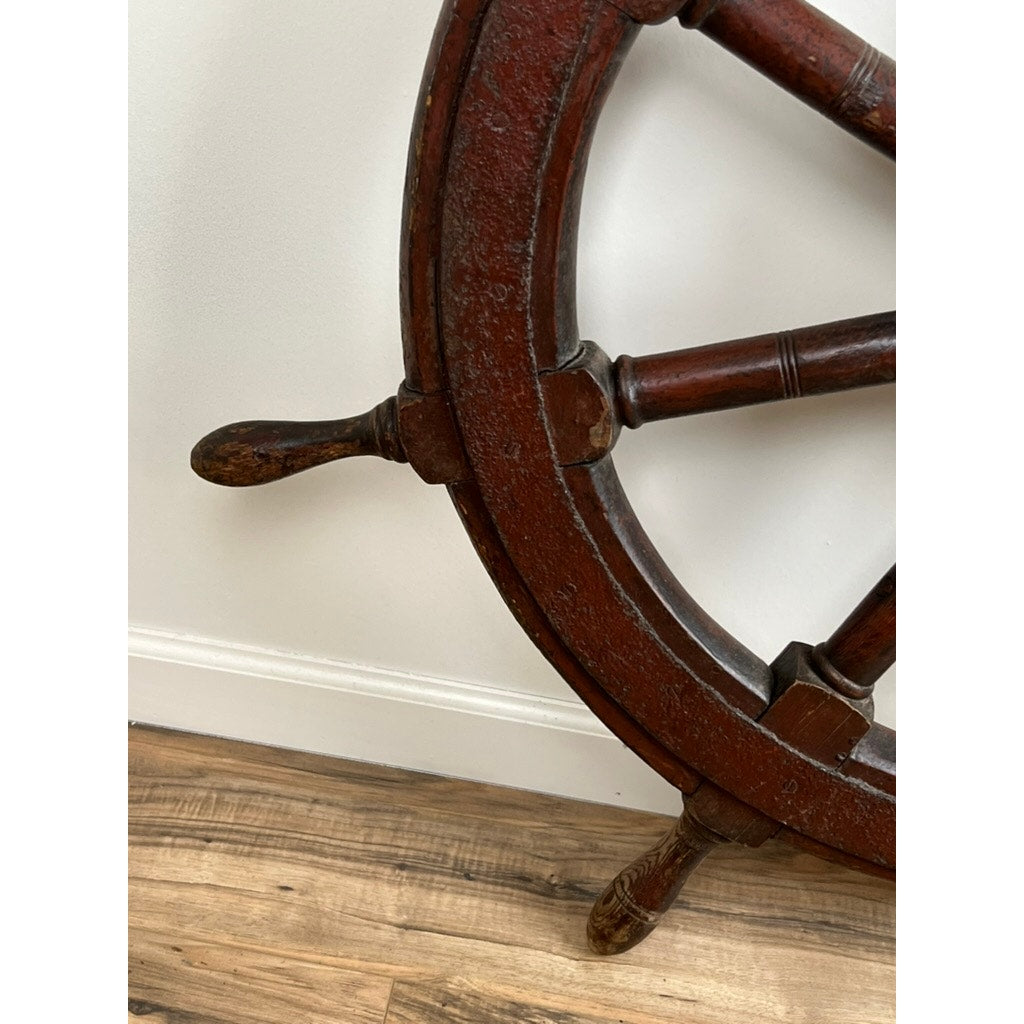 Close-up of an Antique Eight Spoke Mahogany and Iron Ship’s Wheel stamped “1864 US” rests against a white wall on a wooden floor, highlighting its classic 19th-century maritime charm. Only part of the wheel is visible. Brand unknown.