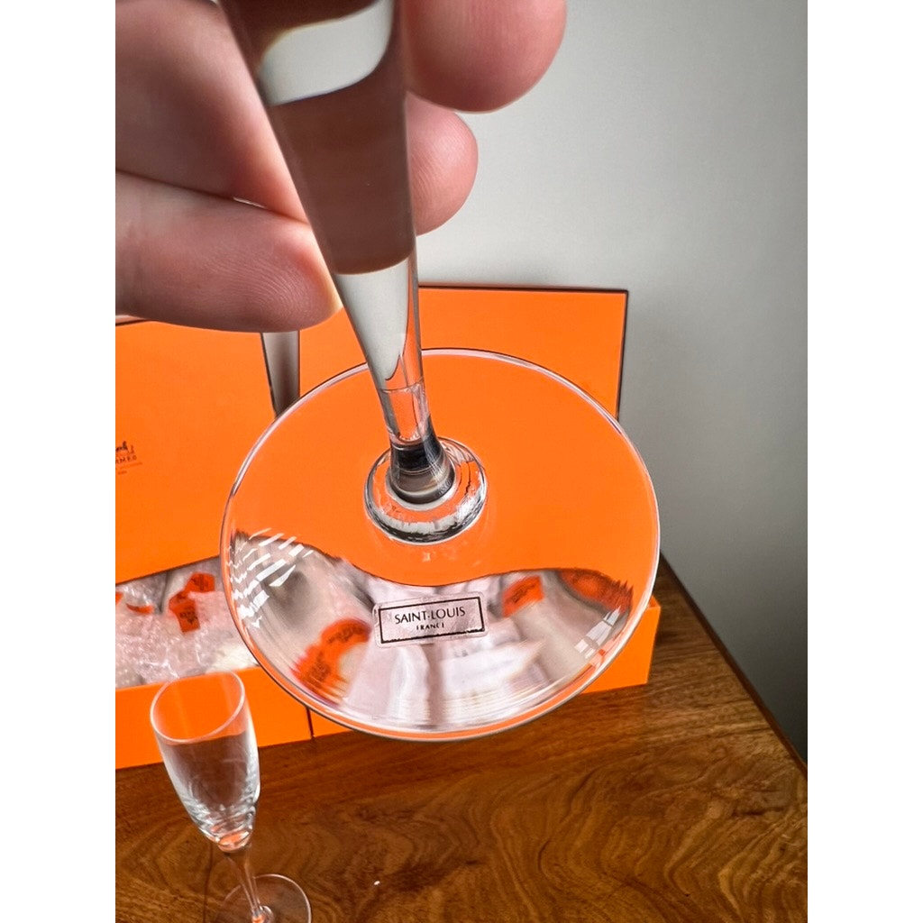 A hand holds a crystal flute above a wooden table with an orange Hermès box—evoking the elegance of Saint-Louis craftsmanship. Another empty flute sits in the background, and the box’s logo is visible through the base of the glass.