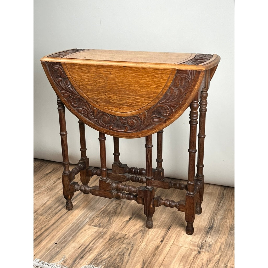 Antique English Oak Carved Gateleg Occasional Table, late 19th century, with floral carvings on the sides, turned legs with crossbar support; displayed on a wood floor against a plain light wall. Brand unknown.