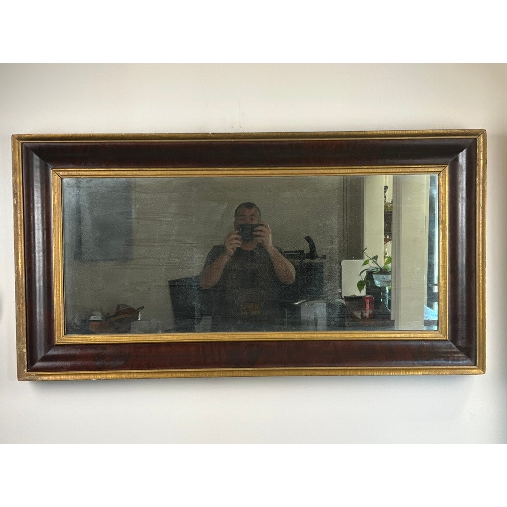 A person photographs their reflection in an Antique American Empire Parcel Gilt Flame Mahogany Ogee Wall Mirror (mid-19th century) with a dark frame and gold trim, mounted on a white wall; household items and plants appear in the background.