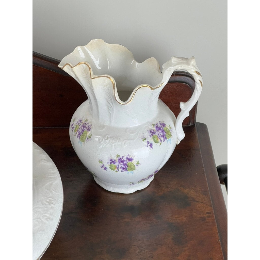 The Antique Victorian Porcelain Wash Stand Set by Leland, c. late 19th century, features a white pitcher with scalloped rim, ornate handle, gold trim and purple flowers on a dark walnut stand with a matching plate—an elegant Victorian accent.