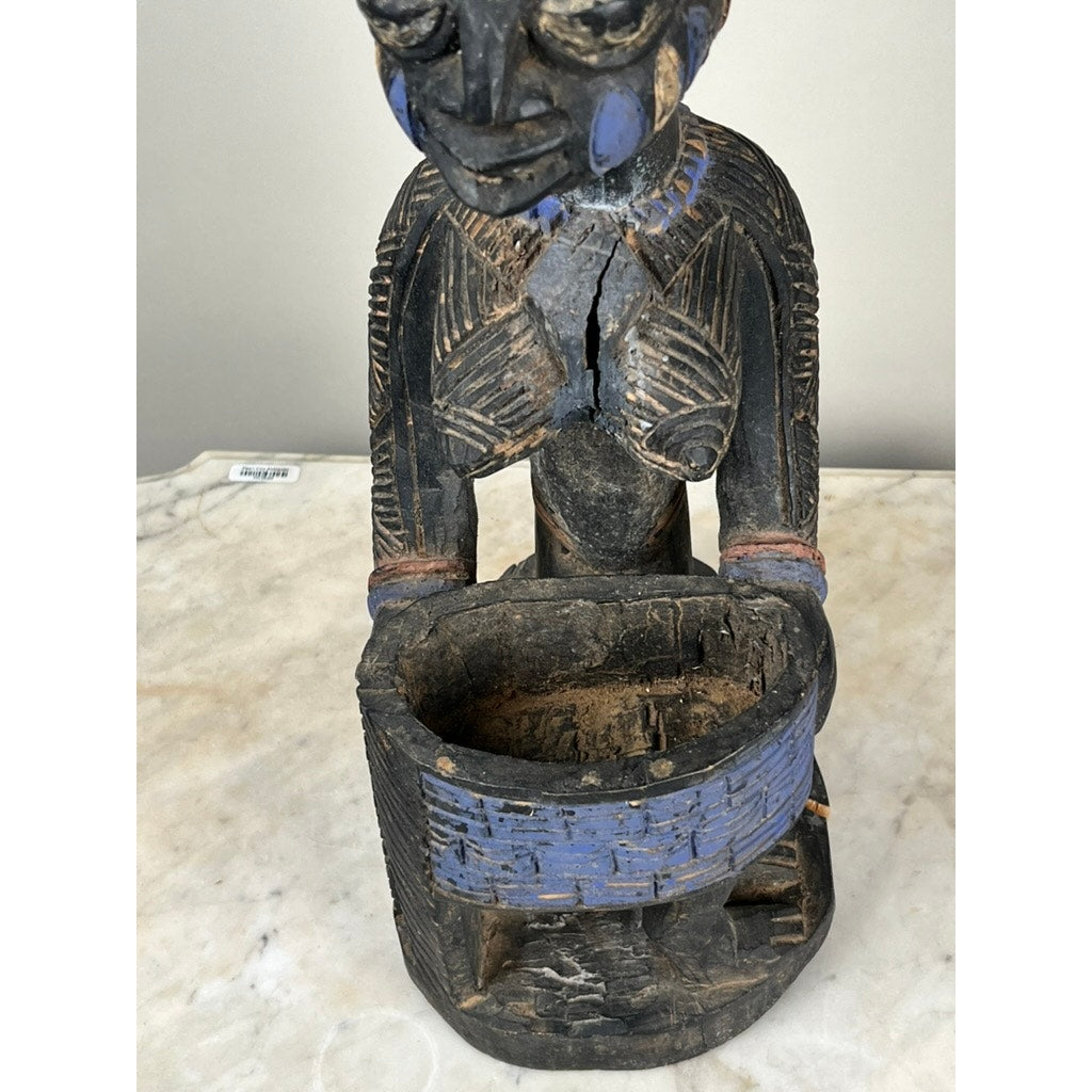 The Antique Igbo Ikenga Shrine Figure, probably from Nigeria, features intricate patterns, visible cracks and wear, and blue accents. It holds a round blue bowl and is displayed on a marble surface. Brand: None or unknown.