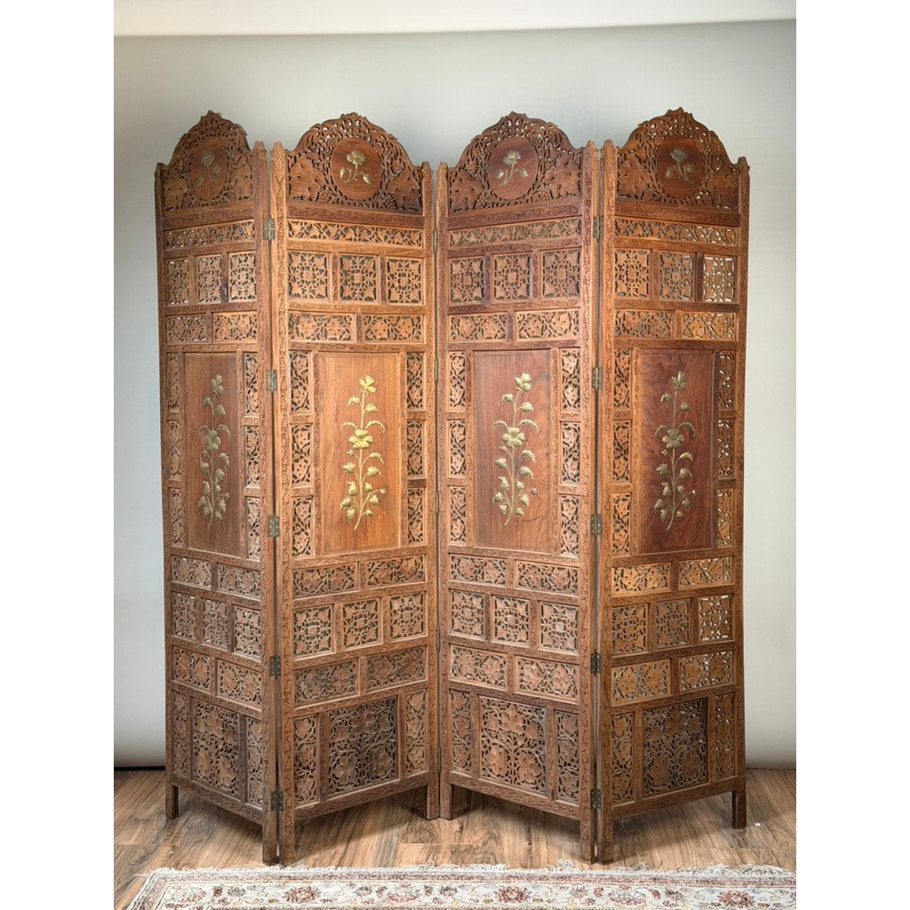 The Early 20th Century Anglo-Indian Carved Teak Four-Panel Screen with Applied Brass Floral Decoration by Pearl City Antiques features intricate carvings, ornate brass floral inlays, and decorative arches crowning each panel. Positioned on a wooden floor with a glimpse of a patterned rug nearby, this antique room divider embodies classic elegance and refined craftsmanship.