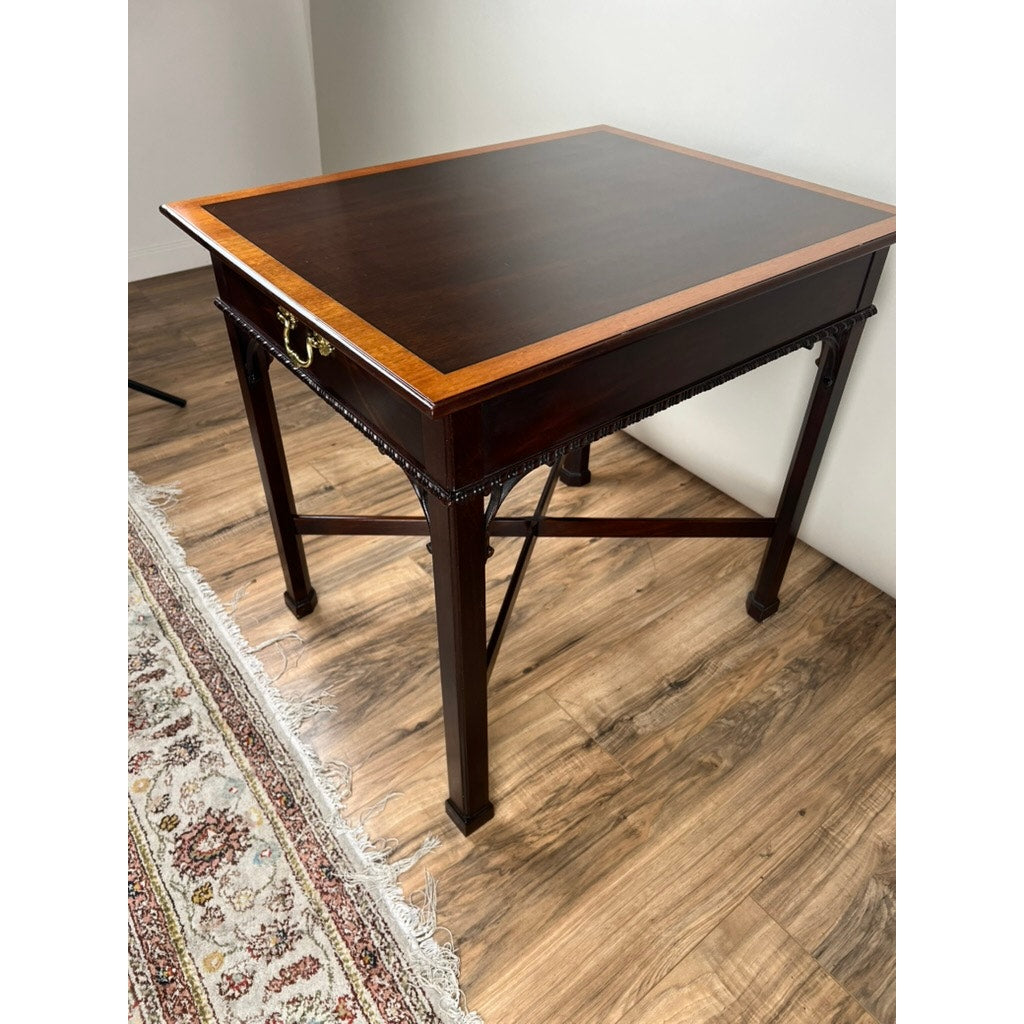 The Stickley Vintage Federal Style X Stretcher One Drawer End Table, featuring a dark wood finish, lighter top border, metal side handle, and cross braces, stands on a wood floor next to a patterned rug.