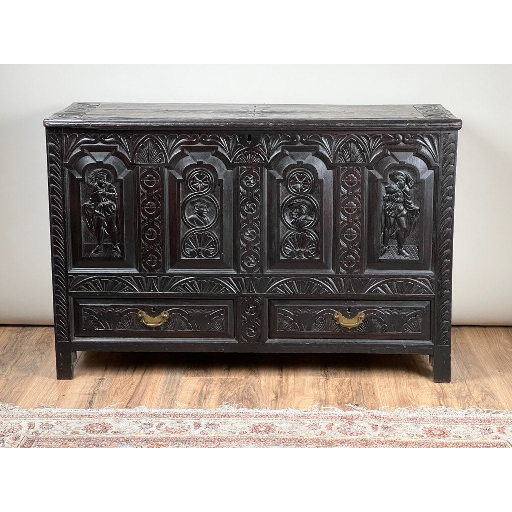 An Antique 18th Century Charles II Style Mule Chest, possibly Irish or Scottish, featuring ornate figure carvings, decorative patterns, two large front panels, and two drawers with brass handles, stands on a wooden floor beside a patterned rug.
