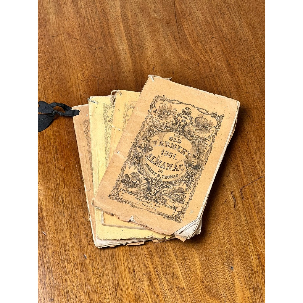 A set of four antique Old Farmer’s Almanacs from 1858, 1861, 1864, and 1865 by Robert B. Thomas features worn, yellowed covers and classic early American print, displayed on a wooden surface. Brand unknown.