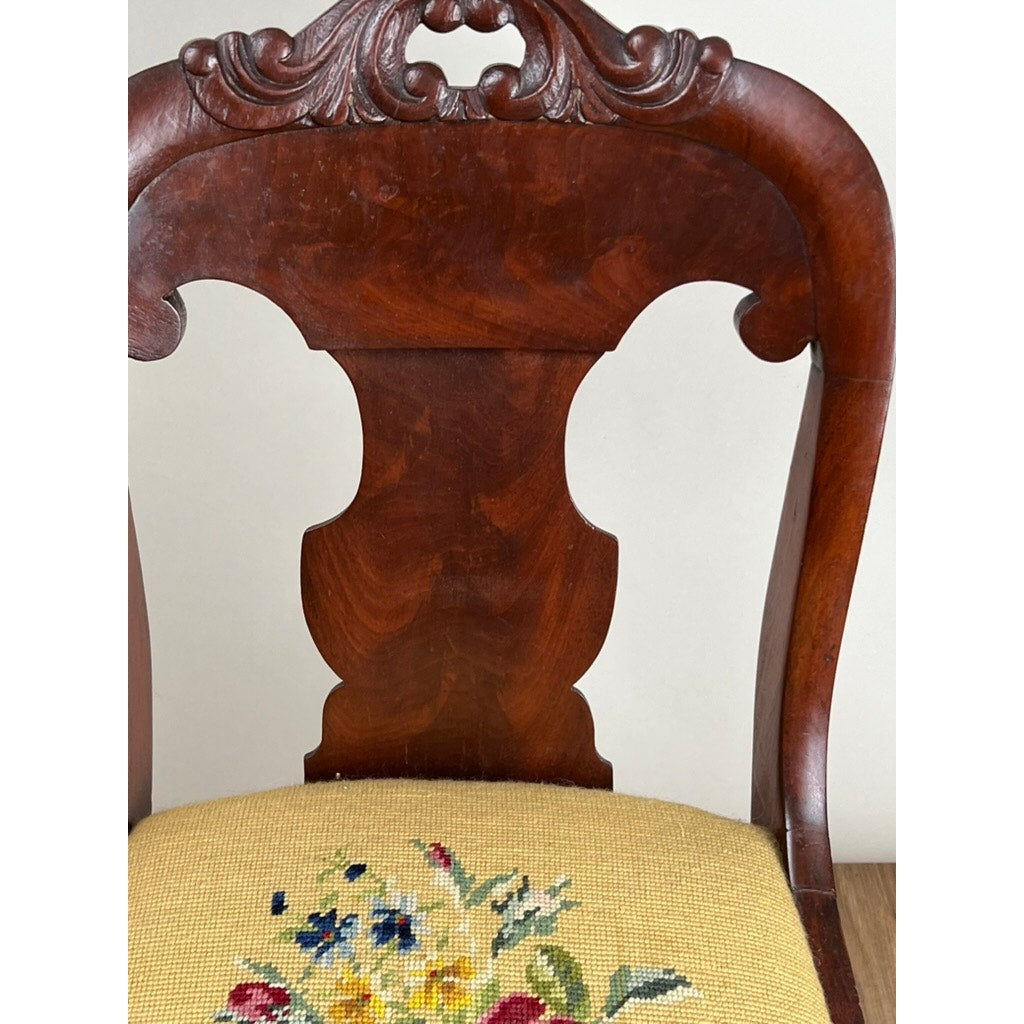 Antique American Empire flame mahogany saber leg side chair, mid-19th century, with a carved backrest and cushioned seat upholstered in colorful antique needlepoint over beige fabric.
