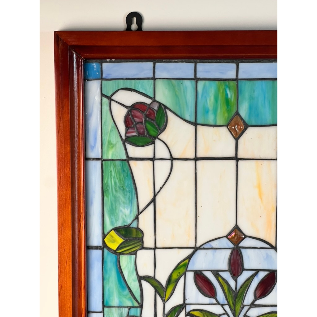 A close-up of the top left corner of a Vintage Stained Glass Wall Hanging 32x20 with a wooden frame, featuring Art Nouveau-style red, yellow, and green floral patterns on blue, white, and beige glass. A wall hook is visible above the frame.