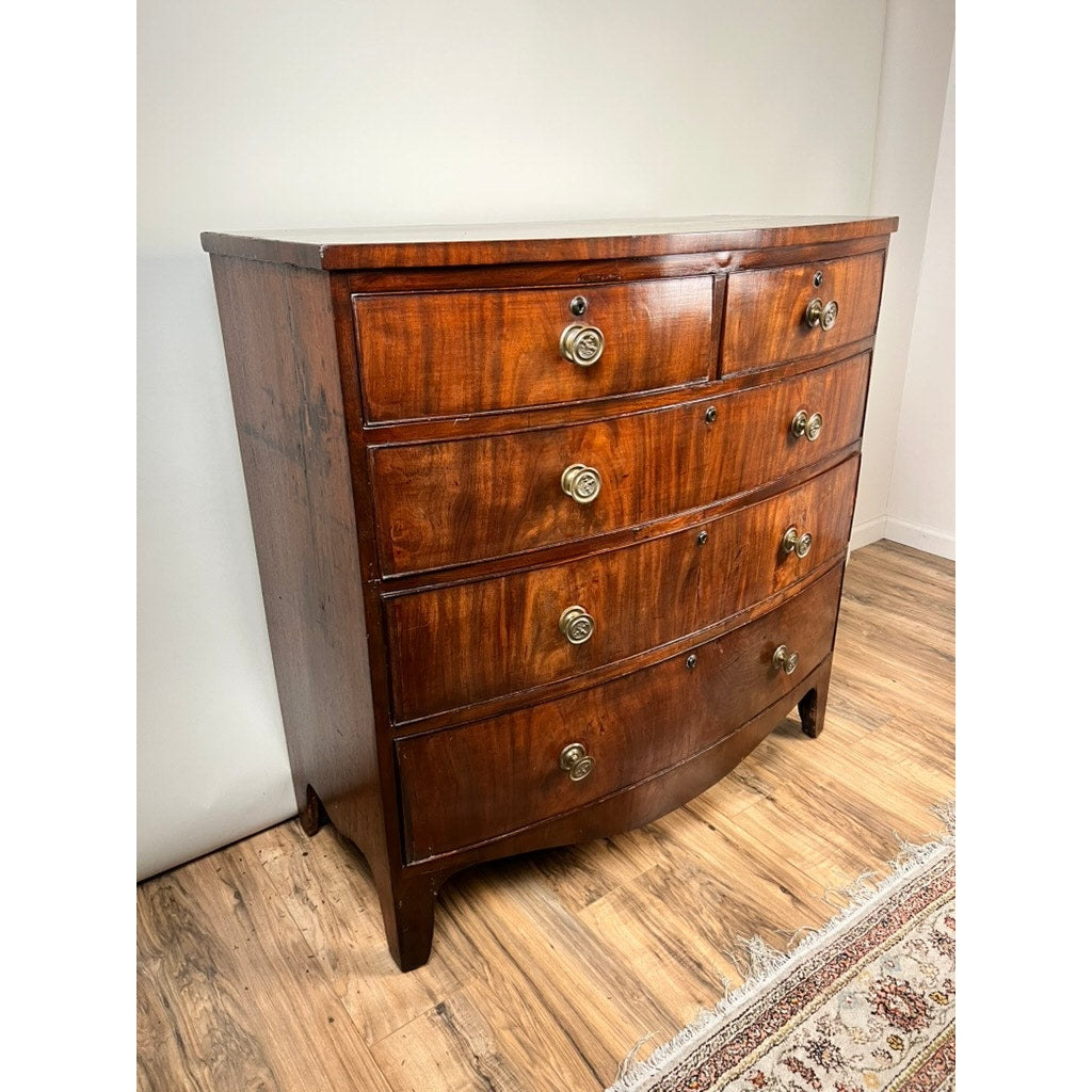 An Antique George III Mahogany Bowfront Chest on French Feet from the late 18th century stands on short legs, featuring four curved drawers with brass handles, placed on a wooden floor next to a patterned rug and white wall.