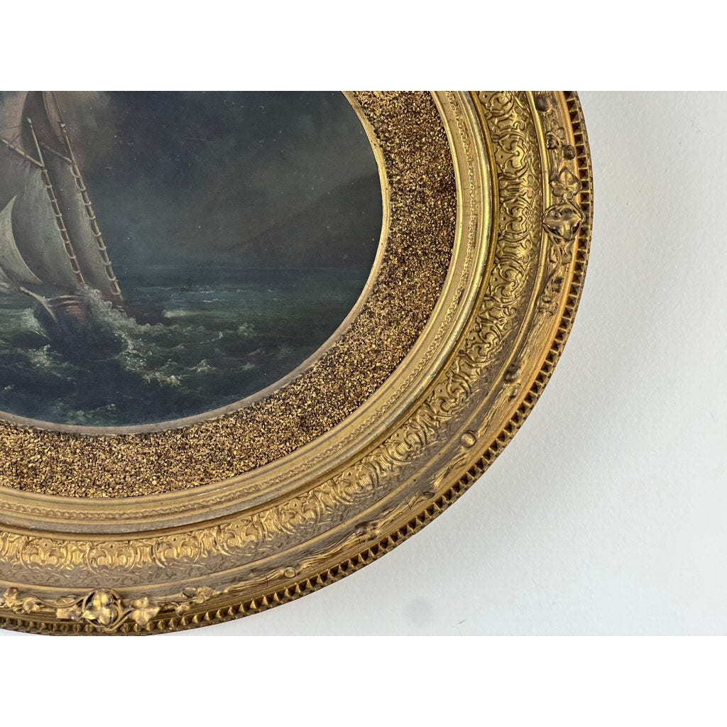 Antique Oval “Storm Tossed Ship” Oil on Board Painting (late 19th c.) in an ornate gilt frame with intricate detailing, depicting a maritime vessel on stormy waters. The elegant piece rests against a plain white wall. Brand: Unknown.