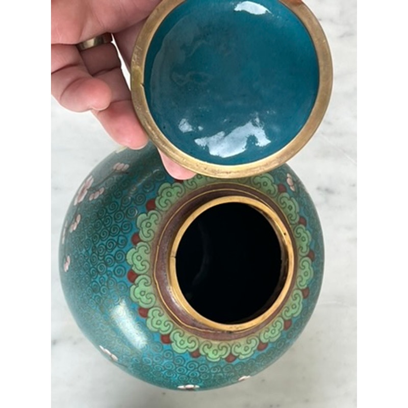 A hand lifts the lid of a Vintage Pair of Chinese Cloisonné Ginger Jars, revealing a dark interior. The jars feature intricate green, blue, and pink floral patterns and rest on a light marble surface as elegant decorative accents.