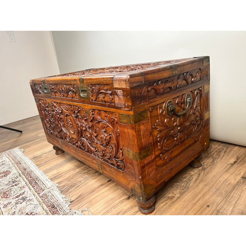 A Chinese Export Carved Camphor Wood Dowry Chest, featuring intricate floral carvings, brass handles, and corner accents, sits on a wooden floor beside a patterned rug against a plain light-colored wall.