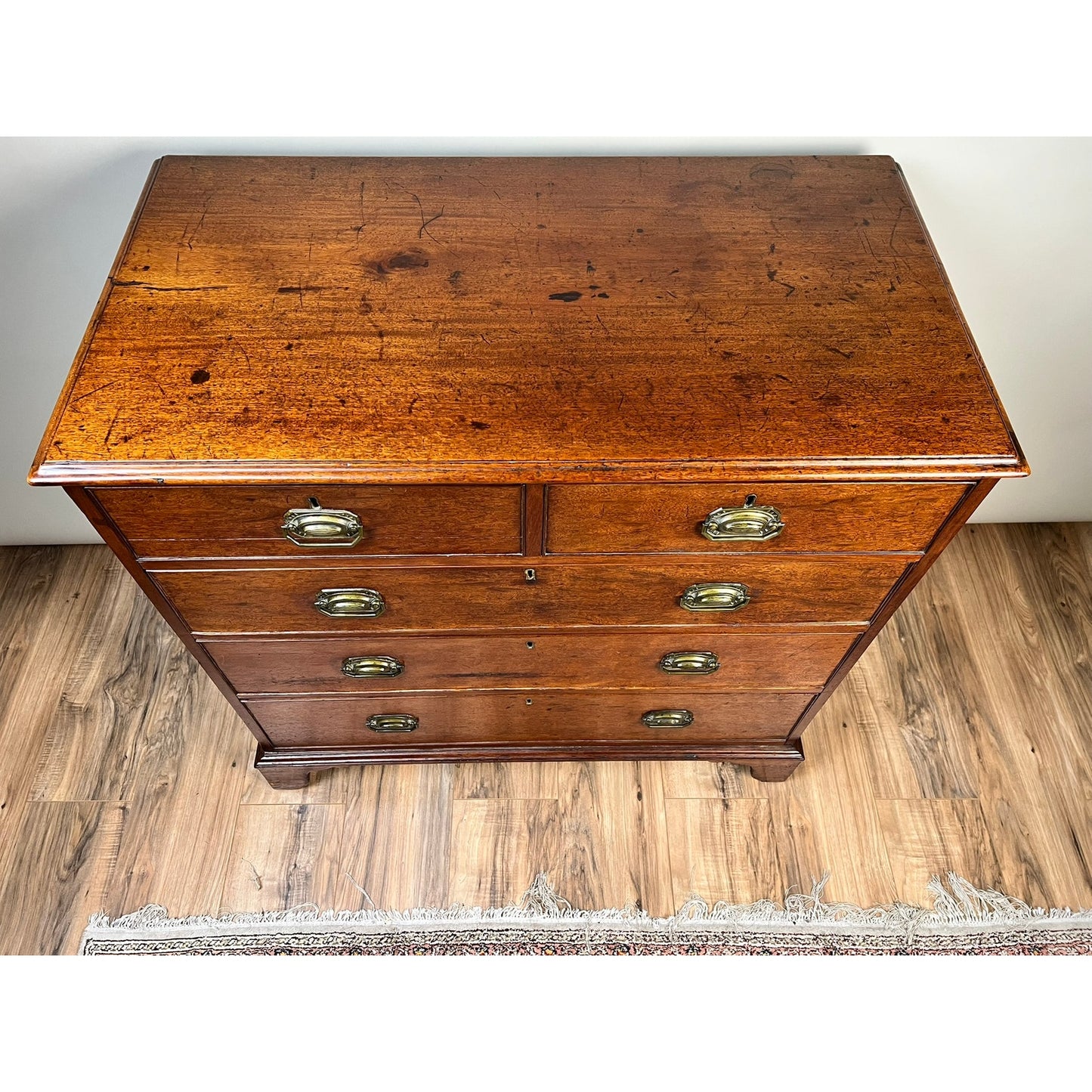 A stunning Late Georgian English Mahogany Chest of Drawers, dating from circa 1780–1810, stands on a wooden floor beside a fringed rug. Featuring four spacious drawers with brass handles and a gently aged surface, this antique piece exemplifies timeless elegance and craftsmanship.