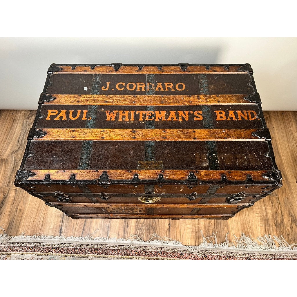 The CA Taylor C.A. Taylor Touring Trunk of John B. Cordaro, Paul Whiteman Orchestra reed player from the 1930s features metal reinforcements and painted labels, shown on a wooden floor next to a patterned rug.