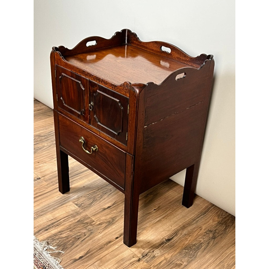 An Antique George III Bedside Commode with pull-out leather surface, late 18th century, features decorative edges, two small doors, brass handles, a single drawer, and four legs, shown against a white wall and wooden floor.
