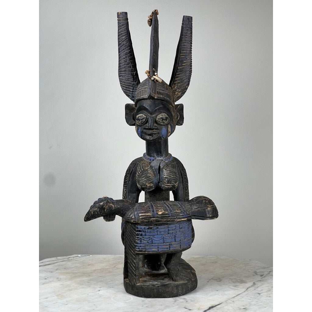 The Antique Igbo Ikenga Shrine Figure, probably from Nigeria, is a dark wooden sculpture seated with tall horn-like protrusions and holding a drum. Detailed features and body markings highlight Igbo artistry. Displayed on white marble against a plain background.