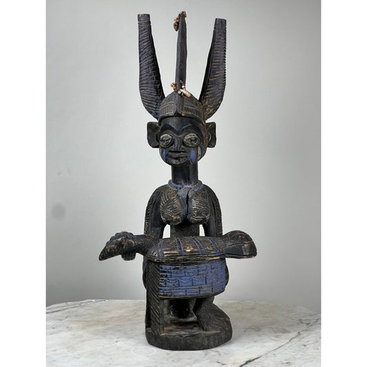 The Antique Igbo Ikenga Shrine Figure, probably from Nigeria and offered by Pearl City Antiques, is a carved wooden statue in the style of West African tribal art. It features tall, curved horns, beaded adornments, and intricate detailing; the figure is seated and holding a drum, displayed on a marble-like surface against a plain background.