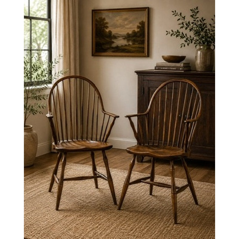 A pair of signed L.E. Partridge Connecticut bench-made continuous-arm Windsor chairs from the 1960s sit on a woven rug in a cozy room featuring a window, potted plant, framed landscape painting by L.E. Partridge, and a wooden sideboard. Warm light fills the space, creating a welcoming atmosphere.