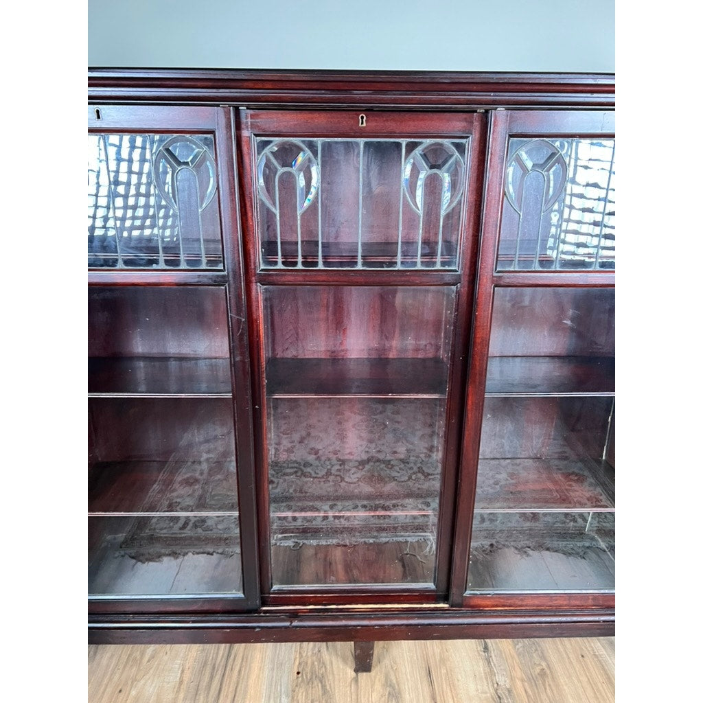 The Antique Arts & Crafts Leaded Glass Mahogany Bookcase c. 1920 features three glass-paneled doors with decorative leaded glass patterns above, empty interior shelves, and stands on a light wooden floor.