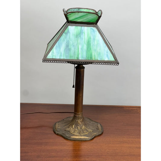 An Antique Arts and Crafts Green Slag Glass Lamp from the 1920s by Pearl City Antiques, featuring a decorative brass base and green slag glass shade, sits on a wooden surface.