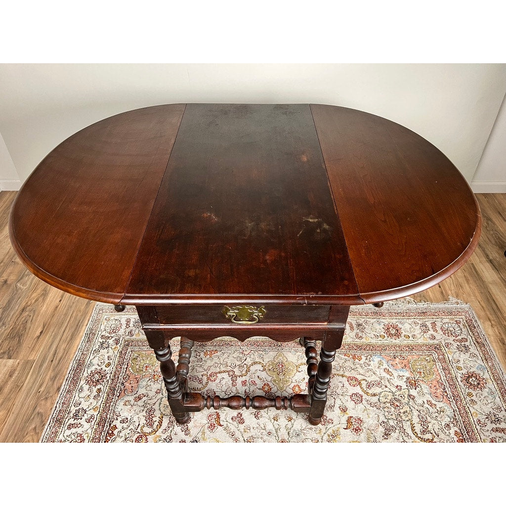 An Antique English William & Mary Walnut One Drawer Gateleg Dining Table, early 18th century, with an oval top and ornate legs, stands on a patterned rug over a wooden floor in a light-colored room. Brand: Unknown.