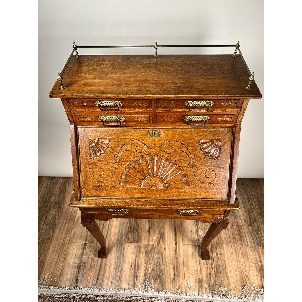 Antique Victorian Oak Ladies Writing Desk c. 1900 featuring ornate carvings, two small drawers, a fold-down front panel with floral and shell designs, and elegant curved legs, displayed on a wood floor. Brand: None/Unknown.