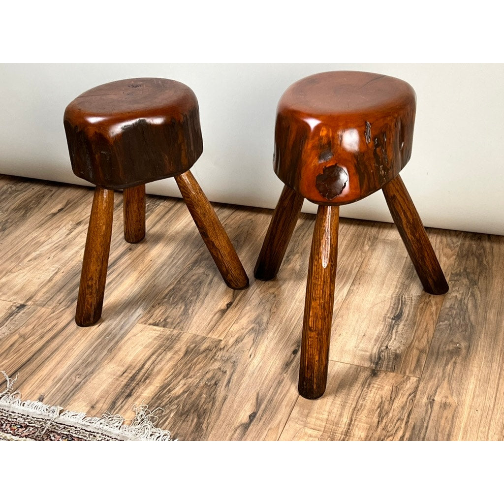 A Pair of vintage, live edge Wabi Sabi cherry stools, signed by Neil Panyik, with round polished seats and three angled legs sit on a wood-patterned floor beside a patterned rug, highlighting their natural grain and unique imperfections.