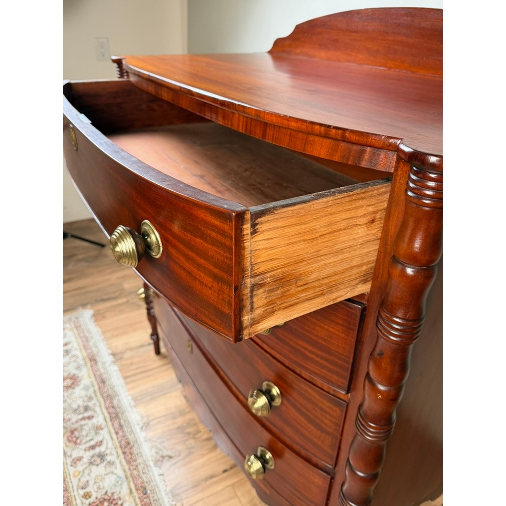 An Early 19th Century New England Federal Mahogany Bow-Front Chest of Drawers with Backsplash, c. 1820–1840, is displayed with its top drawer open to reveal the timeworn interior. This elegant dresser, featuring turned columns and brass knobs typical of its period, stands on a hardwood floor beside a patterned rug.