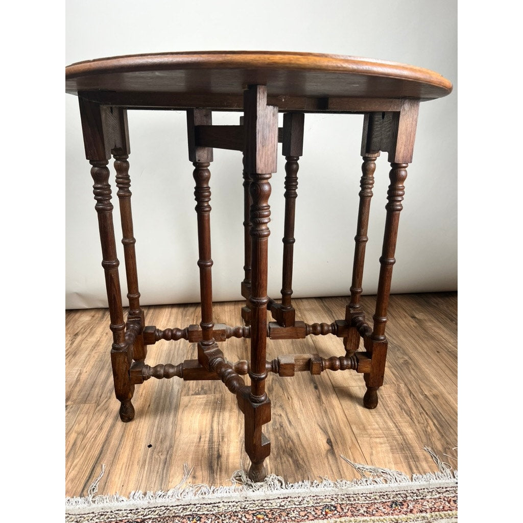 An Antique English Oak Carved Gateleg Occasional Table c. Late 19th Century stands on a wooden floor by a patterned rug, its warm brown finish and intricately turned legs reflecting classic elegance. Brand: None/Unknown.