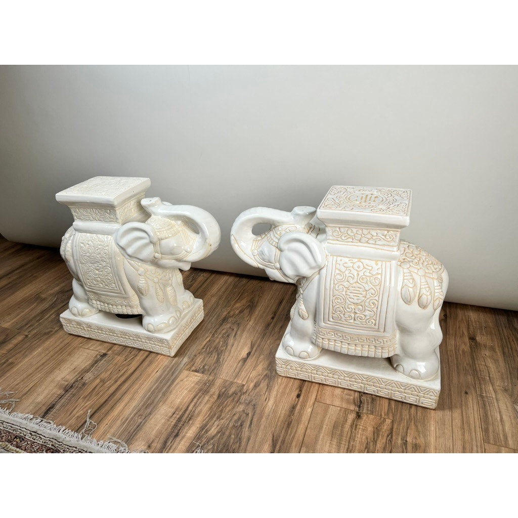 A vintage pair of white-on-white glazed Hollywood Regency style elephant garden seats serve as striking accent tables, placed on a wooden floor against a plain light-colored wall.
