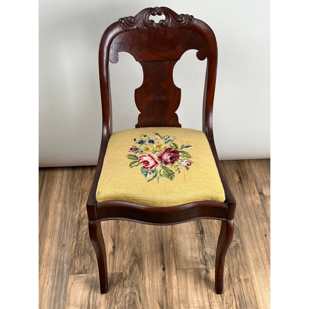 An Antique American Empire Flame Mahogany Saber Leg Side Chair (mid-19th c.) with a carved back, curved legs, and a yellow seat featuring antique floral needlepoint upholstery sits on a wooden floor by a light-colored wall. Brand: None/Unknown.