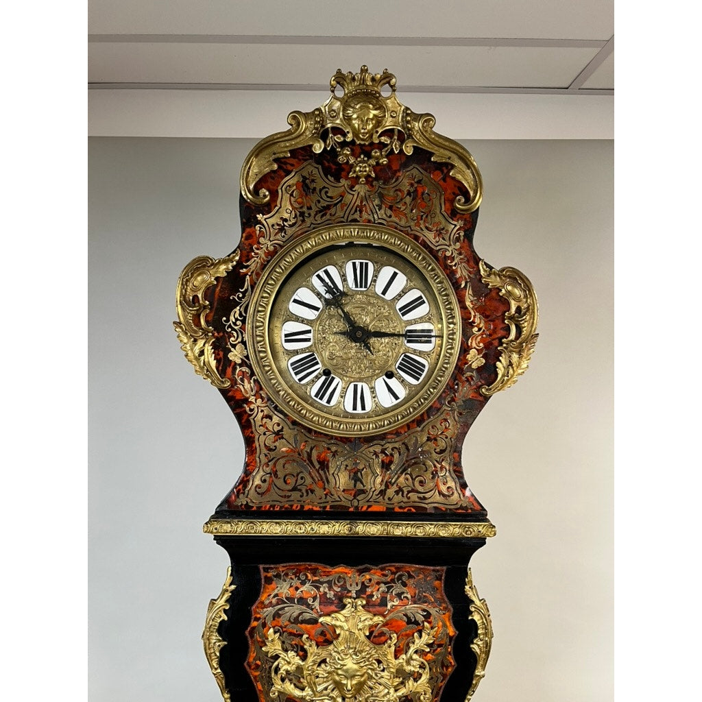 A monumental French Napoleon III Boulle marquetry floor clock with ornate ormolu mounts, dating from circa 1865–1885, features gold detailing, Roman numerals, and elaborate carvings. Set against a plain light-colored wall, this antique timepiece showcases exquisite French decorative artistry reminiscent of Boulle marquetry.