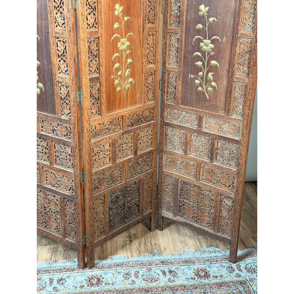 The Early 20th Century Anglo-Indian Carved Teak Four-Panel Screen with Applied Brass Floral Decoration by Pearl City Antiques features intricate carvings and brass floral accents, standing gracefully on a hardwood floor next to a patterned rug.