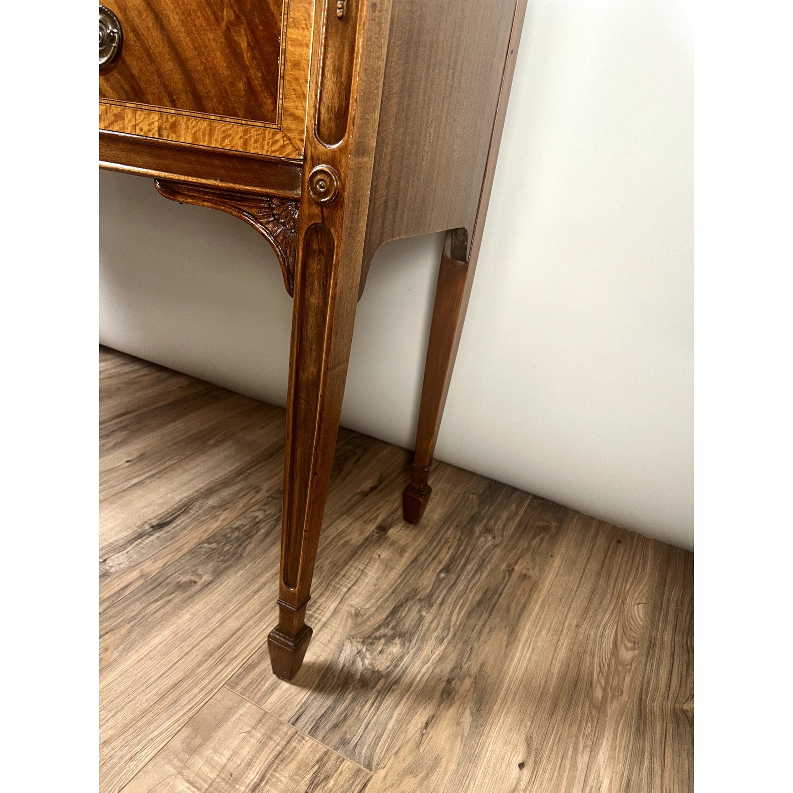 Close-up of the leg and lower corner of a Vintage Federal Style Bow Front Mahogany Sideboard by Paine Furniture Boston, featuring ornate details, standing on a wood-patterned floor against a plain white wall.