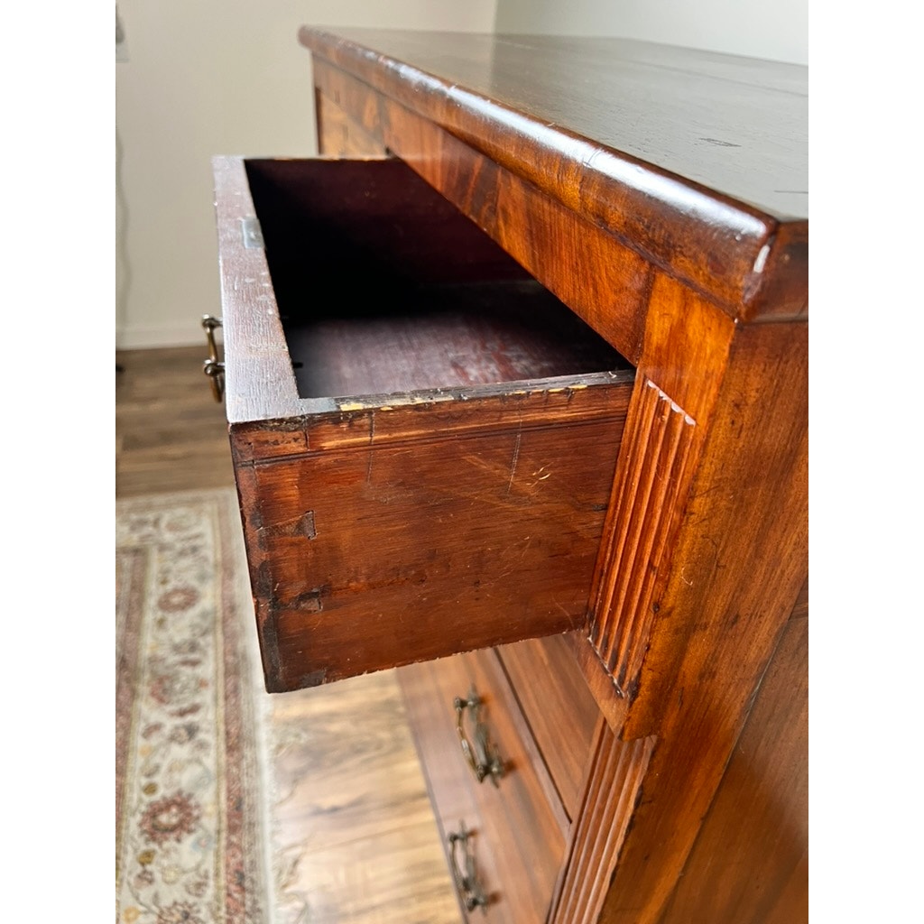 A close-up of an Antique George III Sheraton Flame Mahogany Chest of Drawers (early 19th century) with an open top drawer, revealing its dark interior; the rich brown finish and carved corners sit above a patterned rug and wooden floor.