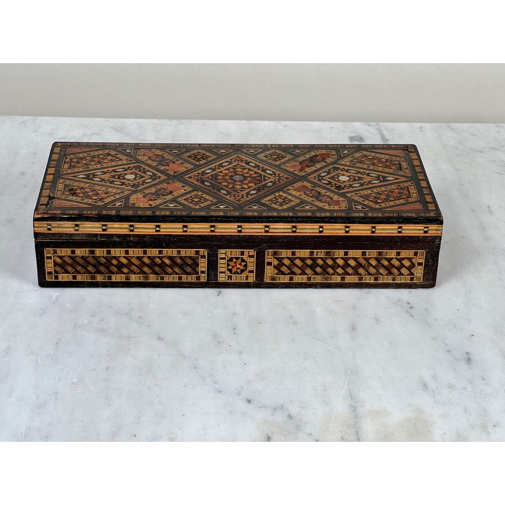 An Antique Persian (likely Syrian) Khatam inlaid wood jewelry box from the early 20th century, adorned with geometric patterns in brown, black, and tan, rests elegantly on a white marble surface.