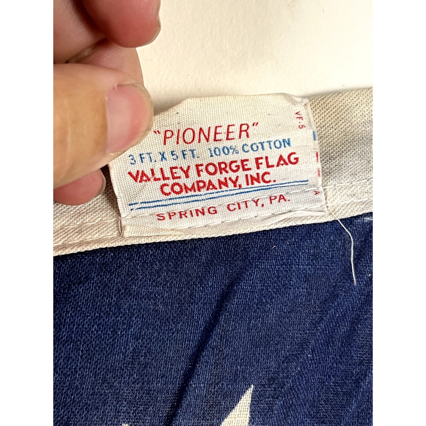 A close-up of a fabric tag reads: Pioneer 3ft x 5ft, 100% cotton, Valley Forge Flag Company, Inc., Spring City, PA. A hand holds one corner of the tag, which is attached to blue fabric featuring a white star—an iconic detail found on the Vintage Valley Forge “76” Bennington American Flag from the Bicentennial Era c. 1976.