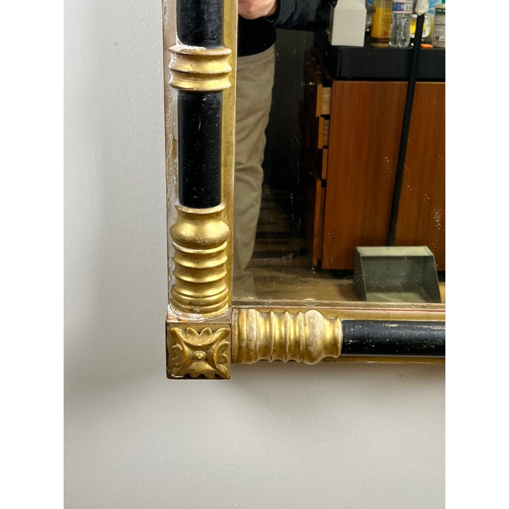 A close-up of an Antique Federal Period Giltwood Mirror with Rosette Corners and Cornucopia c. 1830 reflects part of a person in tan pants and a dark shirt, along with background furniture and objects. Brand: Unknown.
