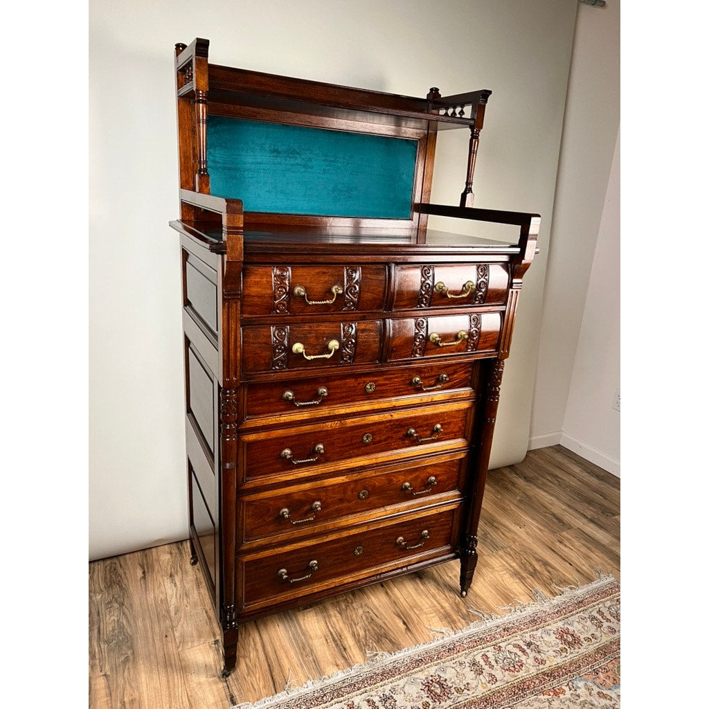 An Antique Victorian Aesthetic Movement Mahogany Four Over Four Gentlemen’s Chest c. 1880 stands on a wood floor by a patterned rug and white walls, featuring ornate drawer handles, carved details, and a blue fabric-lined upper shelf.