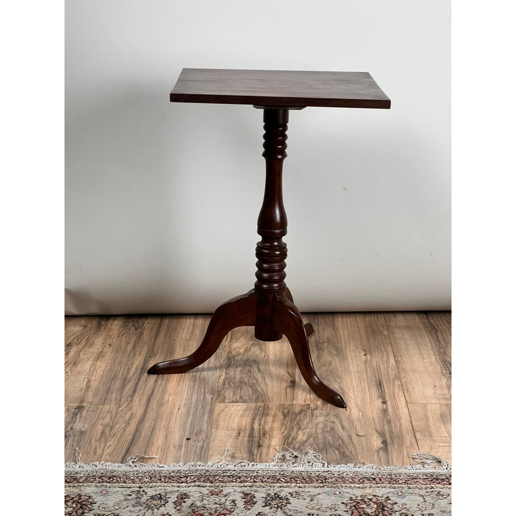 An Antique Low Cherry Candle Stand from the mid-19th century, with a square top, turned central column, and three curved legs, stands on a wooden floor beside part of a patterned rug. Brand: None/Unknown.