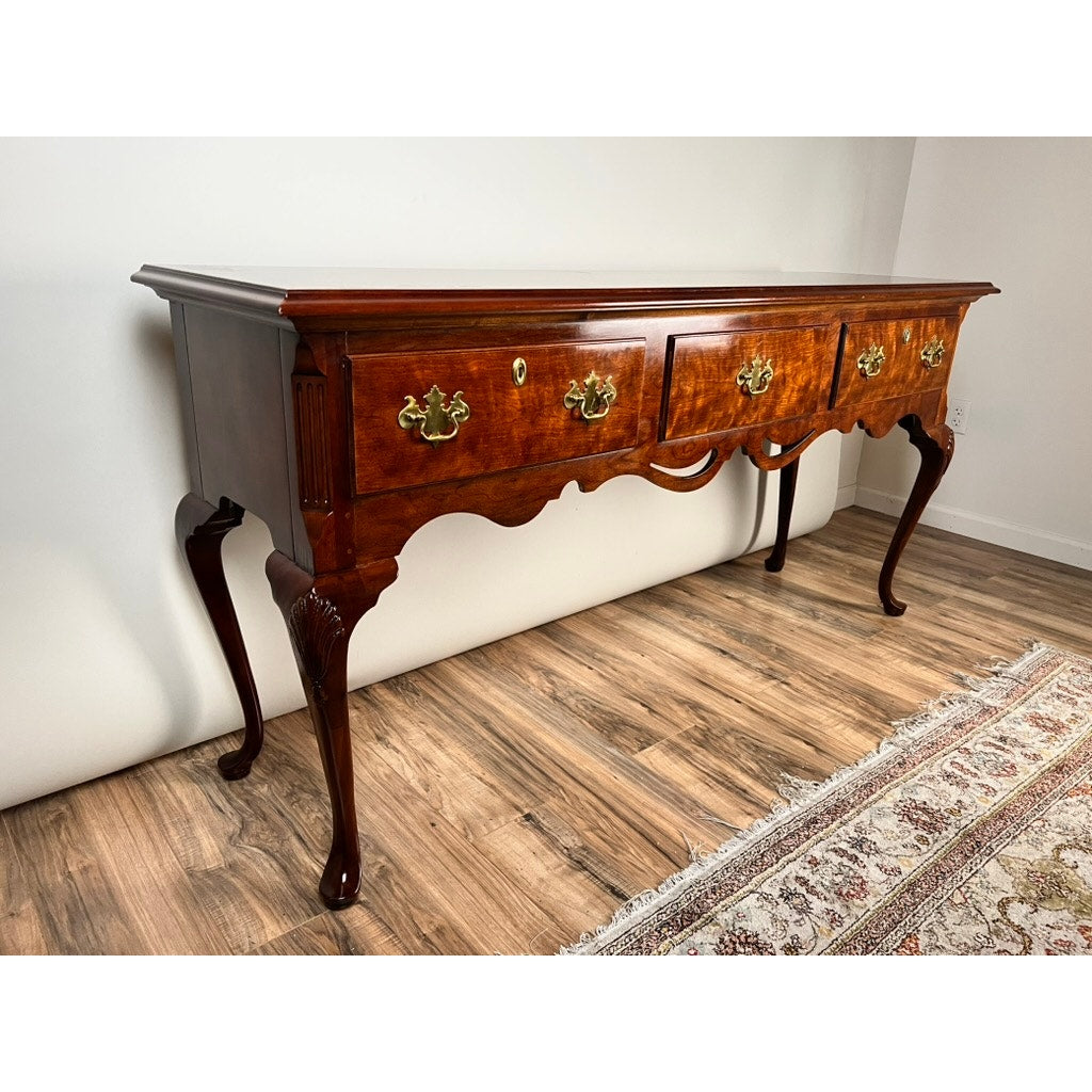 A Vintage Solid Cherry Queen Anne Style Sideboard by Henredon with three drawers, brass handles, cabriole legs, and carved details stands on a wooden floor beside a patterned rug.