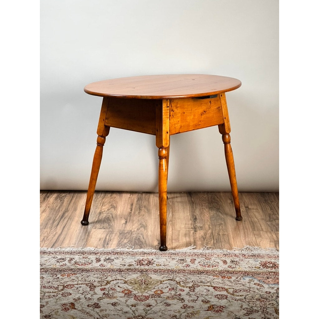 An Antique American Queen Anne Maple Tavern Table, New England, circa 1760–1790, with a round top and four turned legs, features a warm brown finish. It sits on a wood floor next to a patterned rug against a light-colored wall.