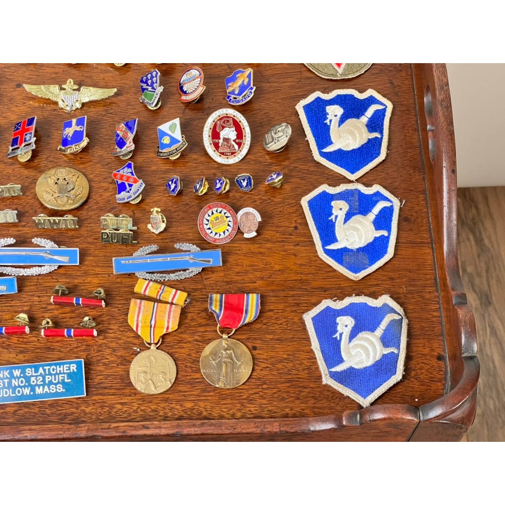 A wooden table displays the Attributed WWII 158th Infantry Bushmasters Grouping of Frank W. Slatcher, Company I, Pacific Theater, with military pins, badges, two medals with ribbons, a blue nameplate, and three blue shield patches featuring a white ram.