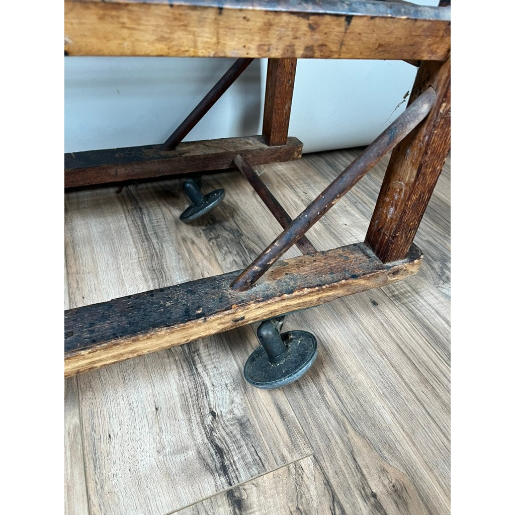 Close-up of the Antique Oak Shoe Factory Rolling Rack on original industrial casters (c. late 19th century), showcasing its aged oak frame and two metal wheels resting on a light wood floor for authentic vintage charm. Brand: Unknown.