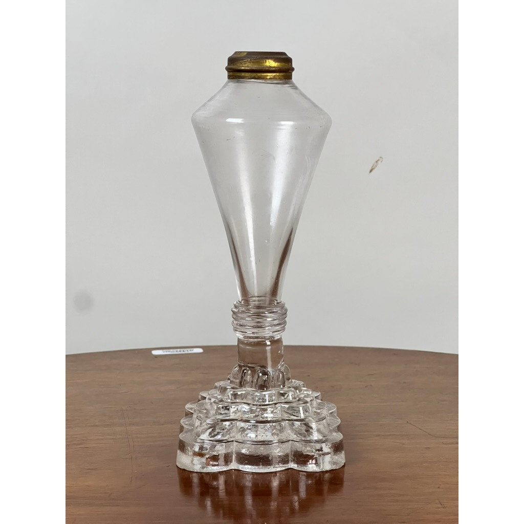 An Attributed Boston & Sandwich Pressed Glass Boudoir Fluid Lamp, Circa 1850, featuring a clear vintage pressed glass body with a faceted stepped base and metal cap, is displayed on a wooden surface against a plain white background.